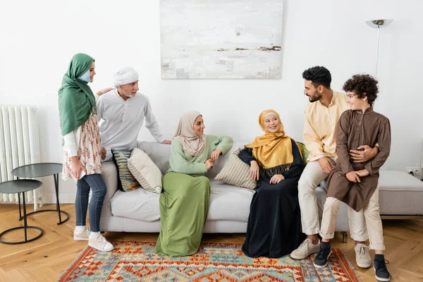 Mujeres interracial feliz sentado en el sofá cerca de la familia musulmana en casa - foto de stock