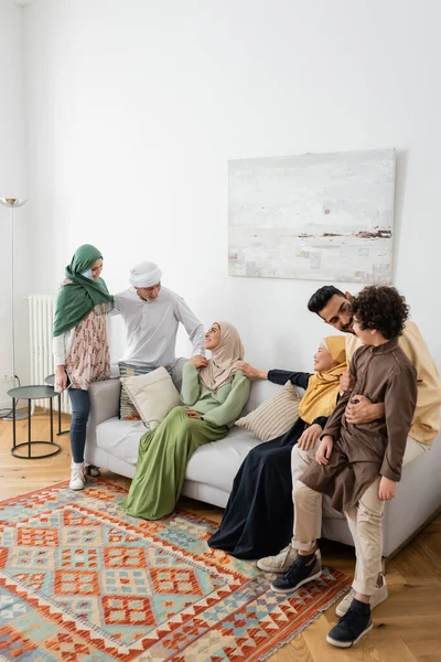 Joven musulmán hombre abrazando árabe hijo cerca alegre multicultural familia en sala de estar - foto de stock