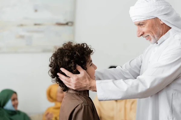 Homme musulman heureux dans turban étreignant la tête du petit-fils arabe près de la famille multiethnique floue — Photo de stock