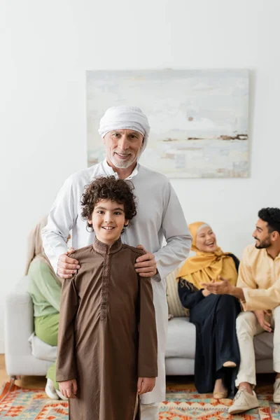 Мусульманський чоловік середнього віку обіймає плече арабського онука біля багатоетнічної родини на розмитому тлі — стокове фото