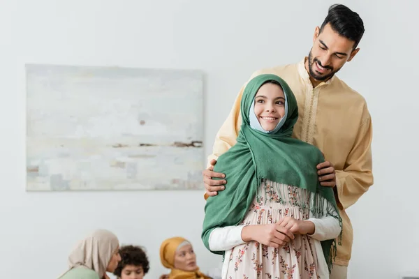 Glücklicher arabischer Mann umarmt muslimische Tochter nahe verschwommener multiethnischer Familie zu Hause — Stockfoto