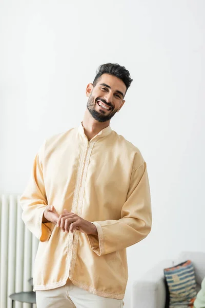 Cheerful arabian man in traditional muslim clothes looking at camera at home — Stock Photo