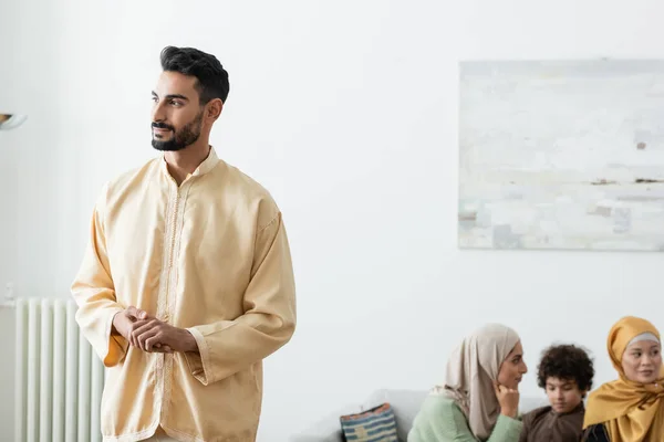Jeune homme musulman regardant loin tout en se tenant près de la famille interracial sur fond flou — Photo de stock