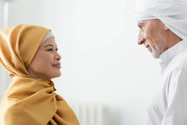 Вид сбоку межрасовой мусульманской пары в традиционной одежде, улыбающейся друг другу дома — стоковое фото