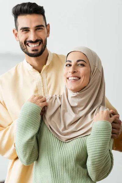 Homme arabe joyeux étreignant jeune femme dans le hijab tout en regardant la caméra — Photo de stock