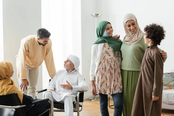 Hombre maduro hablando con hijo adulto cerca de la familia musulmana multiétnica en casa - foto de stock