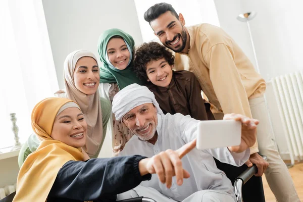 Fröhliche asiatische Frau zeigt mit dem Finger in der Nähe muslimischer Mann macht Selfie mit interrassischen Familie — Stockfoto