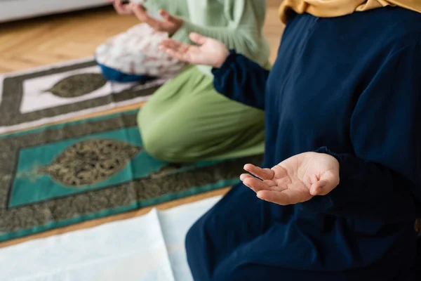 Cropped view of blurred muslim women in traditional clothes praying on rugs at home — Stock Photo