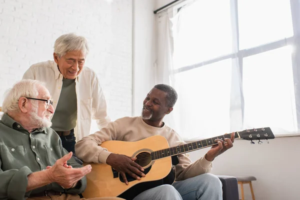 Sonriente hombre afroamericano tocando la guitarra acústica cerca de amigos interracial en casa - foto de stock