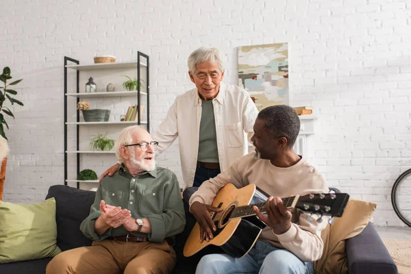 Hombres multiétnicos positivos mirando a un amigo afroamericano tocando la guitarra acústica en casa - foto de stock