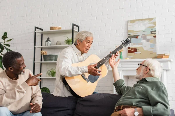 Asiatico uomo holding acustica chitarra vicino allegro multietnico amici a casa — Foto stock