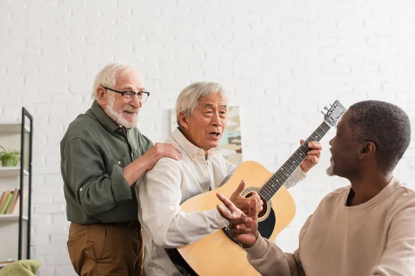 Позитивный старший мужчина обнимает азиатского друга с акустической гитарой дома — стоковое фото