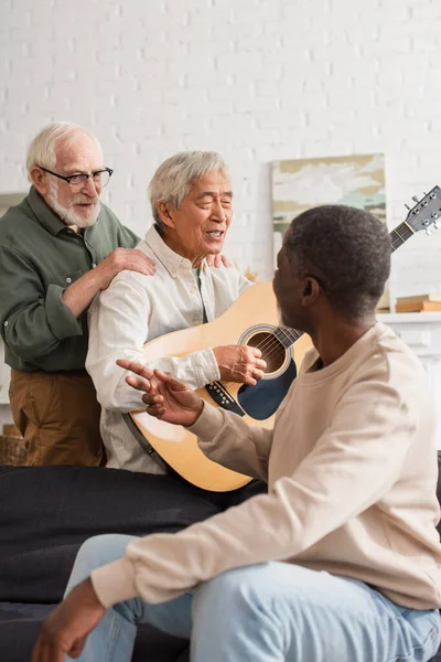 Positivo uomo anziano abbracciare amico asiatico suonare la chitarra acustica in soggiorno — Foto stock