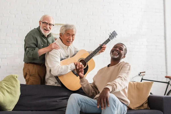 Feliz sénior interracial los hombres con acústica guitarra mirando cámara en casa - foto de stock