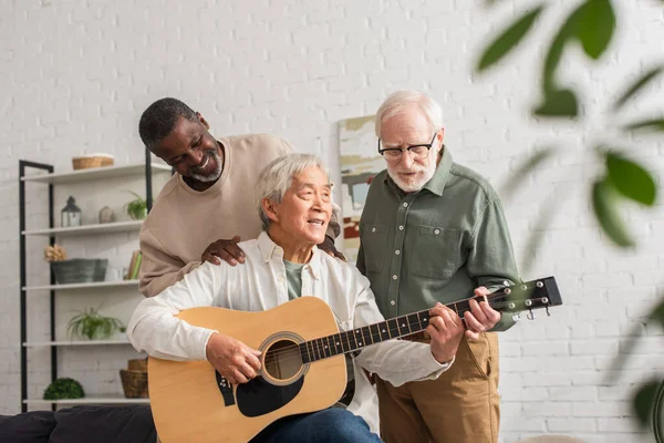 Sorridente africano americano uomo abbracciare asiatico amico giocare acustica chitarra a casa — Foto stock