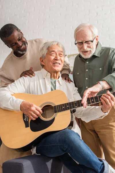 Счастливые старшие мультиэтнические мужчины стоят рядом с азиатским другом, играя на акустической гитаре дома — стоковое фото
