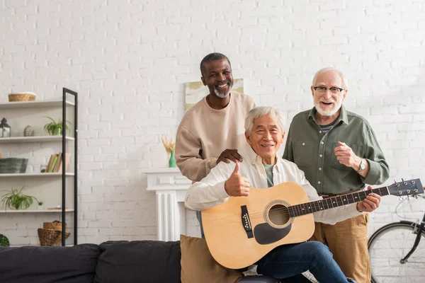 Sorrindo asiático homem segurando guitarra acústica e mostrando como perto interracial amigos em casa — Fotografia de Stock