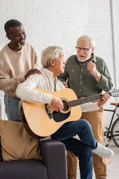 Alegre interracial los hombres de pie cerca asiático amigo jugando acústica guitarra en casa - foto de stock