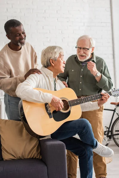 Alegre interracial homens de pé perto ásia amigo jogar acústico guitarra no casa — Fotografia de Stock