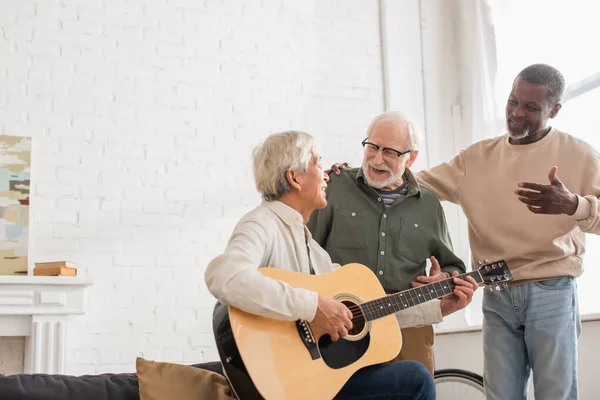 Позитивные старшие друзья смотрят на азиатского мужчину, играющего на акустической гитаре дома — стоковое фото