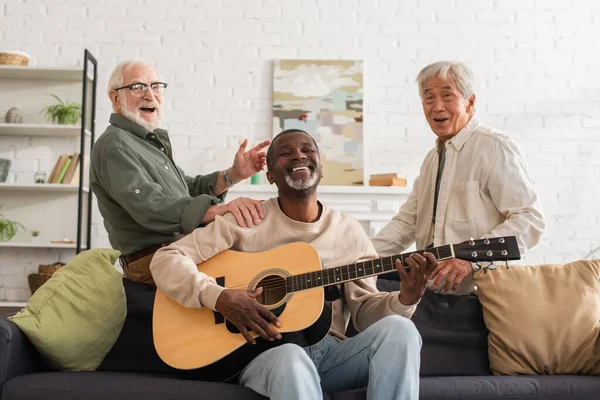 Heureux homme afro-américain jouant de la guitare acoustique près d'amis interraciaux regardant caméra à la maison — Photo de stock