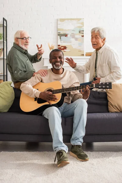 Interracial senior men sing near african american friend playing acoustic guitar at home — Stockfoto