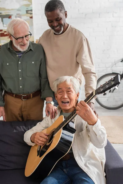 Sénior asiático homem tocando guitarra acústica e cantando perto de amigos multiétnicos em casa — Fotografia de Stock