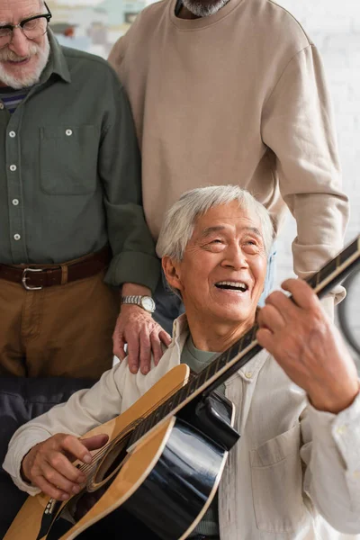 Anziano asiatico uomo giocare acustica chitarra vicino interrazziale amici a casa — Foto stock