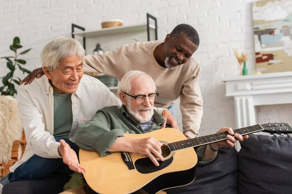 Positivos pensionistas interracial mirando a un amigo tocando la guitarra acústica en casa - foto de stock