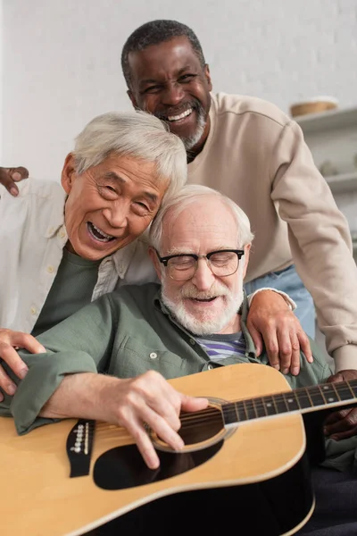Веселые межрасовые старшие друзья играют на акустической гитаре и обнимаются дома — стоковое фото