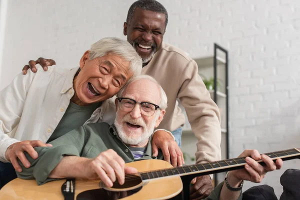 Feliz senior interracial amigos tocando la guitarra acústica y mirando a la cámara en casa - foto de stock