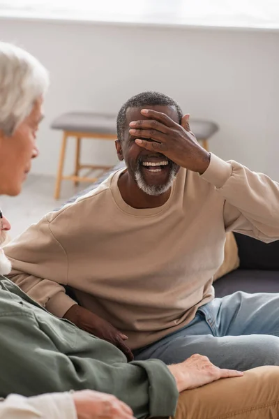 Alegre hombre afroamericano cubriendo la cara cerca de amigos mayores en el sofá - foto de stock