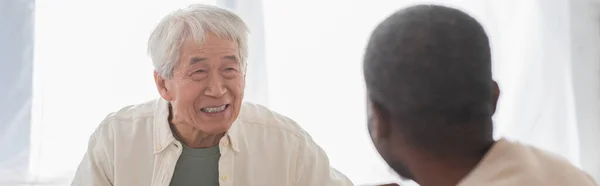 Positivo asiático hombre mirando africano americano amigo en casa, bandera - foto de stock