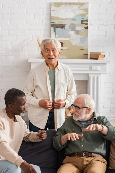 Positivo asiatico uomo guardando fotocamera vicino senior interrazziale amici parlando su divano — Foto stock