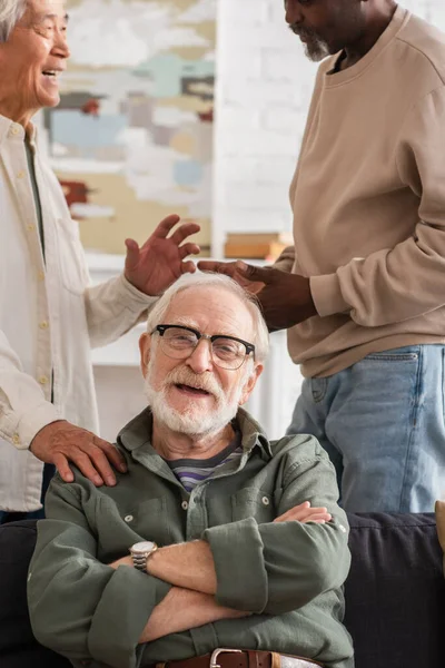 Anciano sonriendo a la cámara cerca de amigos interracial hablando en casa - foto de stock