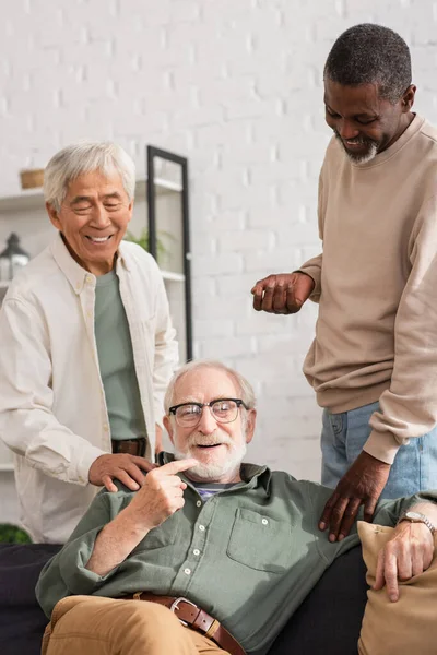 Happy multiethnic men looking at senior friend on couch in living room — Stock Photo