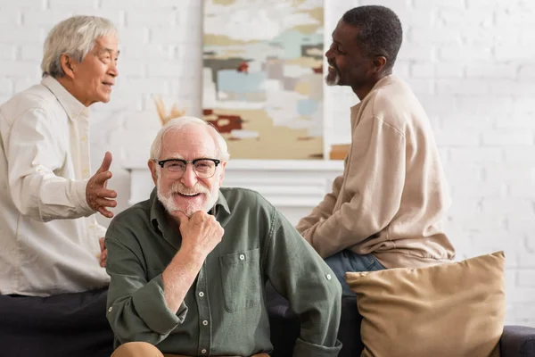 Positive elderly man looking at camera near interracial friends at home — Stock Photo