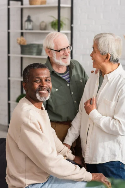 Hombre afroamericano sonriendo a cámara cerca de jubilados multiétnicos en casa - foto de stock