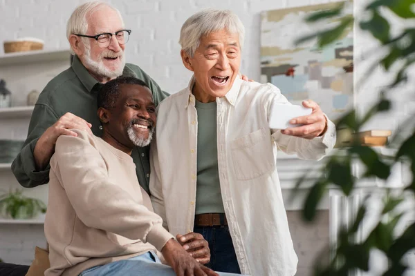 Senior asian man taking selfie near multiethnic friends at home — Stock Photo