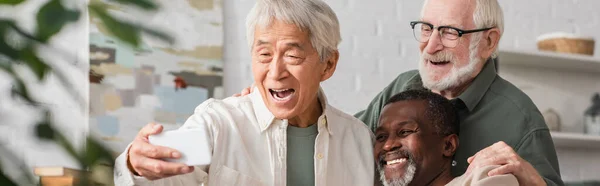 Asian man taking selfie on smartphone with multiethnic friends at home, banner — Stock Photo