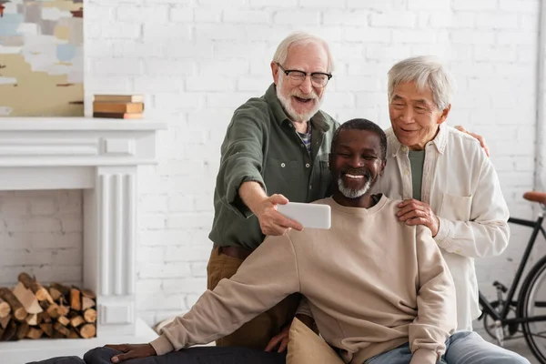 Elderly man taking selfie on smartphone near multiethnic friends at home — Stock Photo