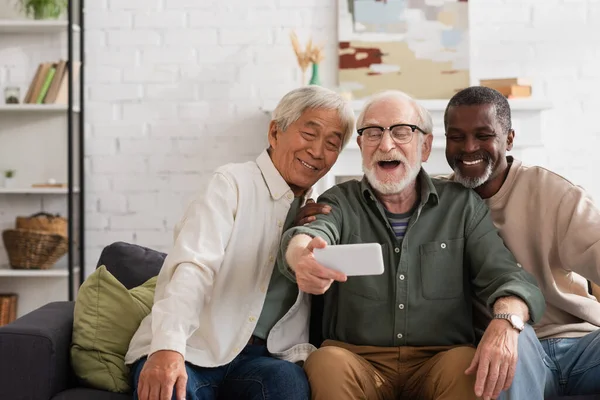 Cheerful multicultural friends taking selfie on smartphone on couch at home — Stock Photo