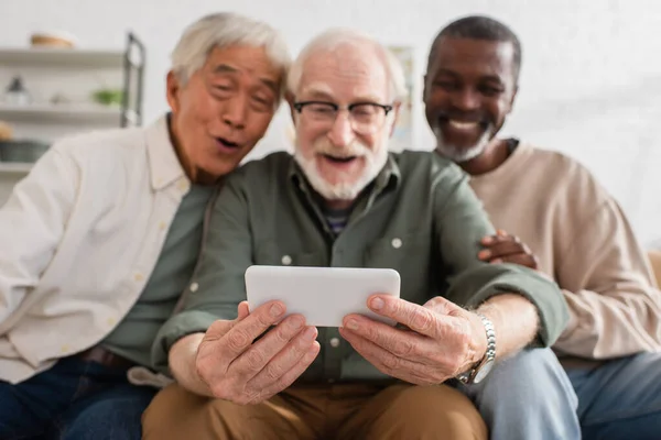 Smartphone en manos de un hombre mayor borroso cerca de amigos interracial en casa — Stock Photo
