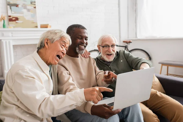 Positive interracial friends pointing at laptop on couch at home — Stock Photo