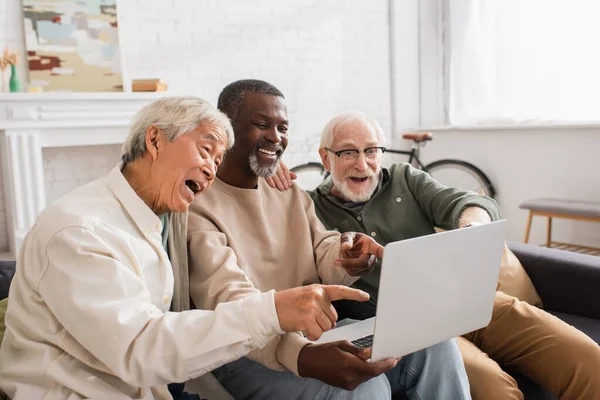 Positivo interracial amigos apontando para laptop no sofá em casa — Fotografia de Stock