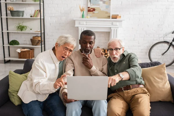 Sorprendidos amigos mayores multiétnicos mirando a la computadora portátil en casa - foto de stock