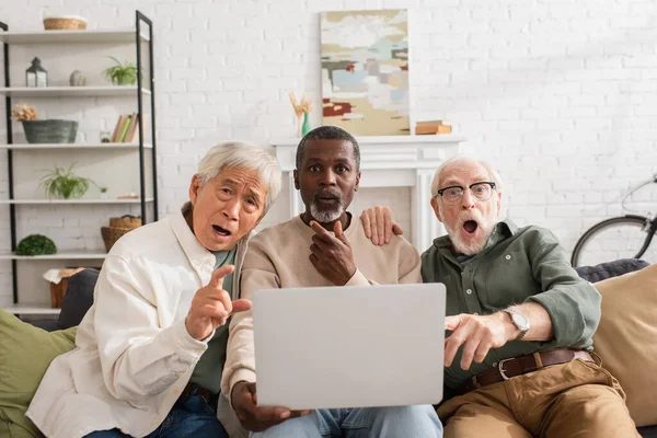 Shocked multicultural friends looking at camera near laptop at home — Stock Photo