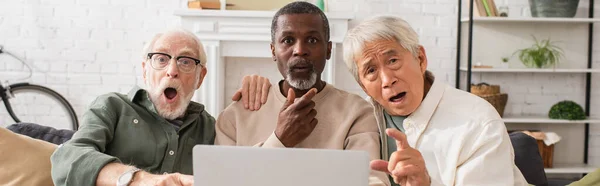 Shocked multicultural friends looking at camera near laptop in living room, banner — Stock Photo