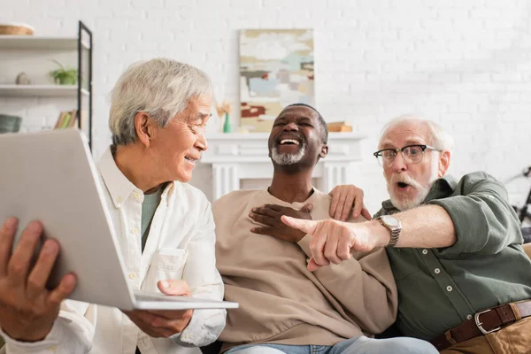 Positive asian man holding laptop near interracial friends at home — Stock Photo