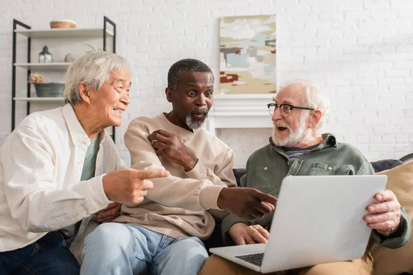 Eccitato uomo afro-americano che punta al computer portatile vicino agli amici multietnici a casa — Foto stock